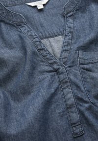 Denimtop med V-ringning, korta ärmar och bröstficka. Tygkvaliteten är mjuk och färgen är en medelblå nyans med subtila blekningar.