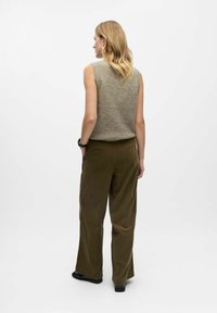 Mouwloos beige gebreid topje, gecombineerd met hoge taille, wijde olijfgroene broek. Soepele textuur met subtiele kleurnuances. Zwarte schoenen.