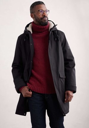 Abrigo negro impermeable con capucha y forro de polar. Suéter de cuello alto de punto rojo, jeans de mezclilla azul. Hombre de pie con las manos en los bolsillos.