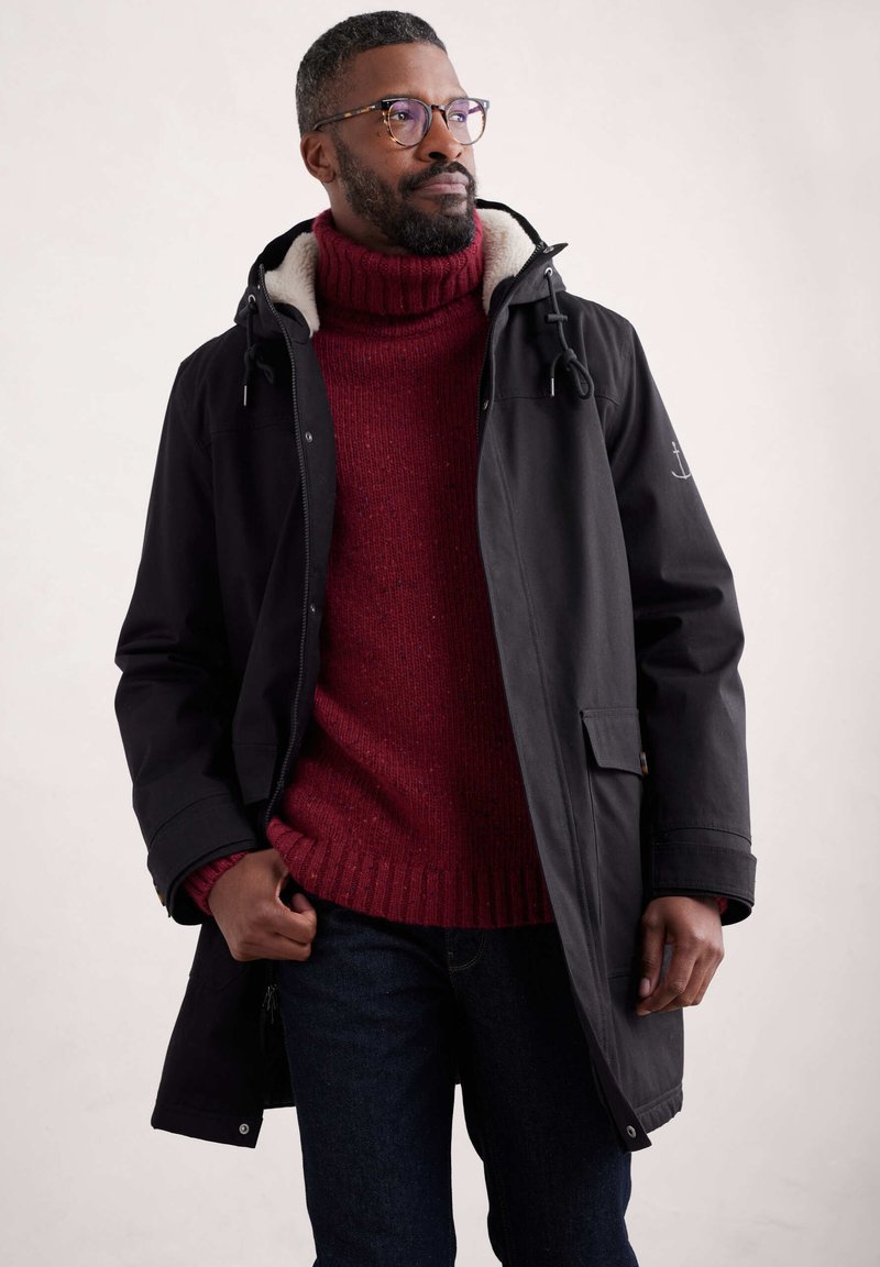 Manteau imperméable noir avec capuche, doublure en polaire. Pull à col roulé tricoté rouge, jean en denim bleu. Homme debout les mains dans les poches.