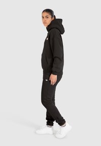 Haut à capuche noir avec poche avant et logo brodé, associé à un jogging noir assorti. Le modèle porte des baskets blanches avec des accents de logo.