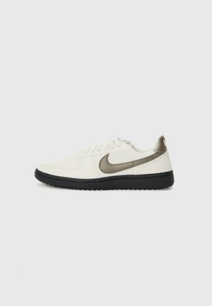 Biały niski sneaker z czarną podeszwą i szarym logo Nike Swoosh z boku, pokazany od zewnętrznej strony na jasnoszarym tle.
