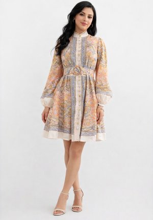 Kobieta stojąca w pastelowej sukience w wzór paisley z długimi bufkami, paskiem w talii i beżowymi sandałkami na kostkę.