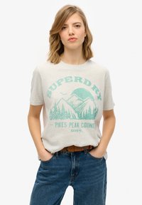Lys grå kortærmet t-shirt med turkis bjerggrafik og tekst. Parret med blå jeans og et brunt bælte, mod en hvid baggrund.