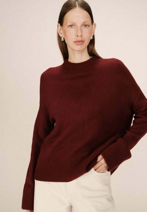 Pull en maille bordeaux avec un col haut et côtelé et des manches longues et larges. Texture douce et coupe décontractée, associé à un pantalon de couleur claire.