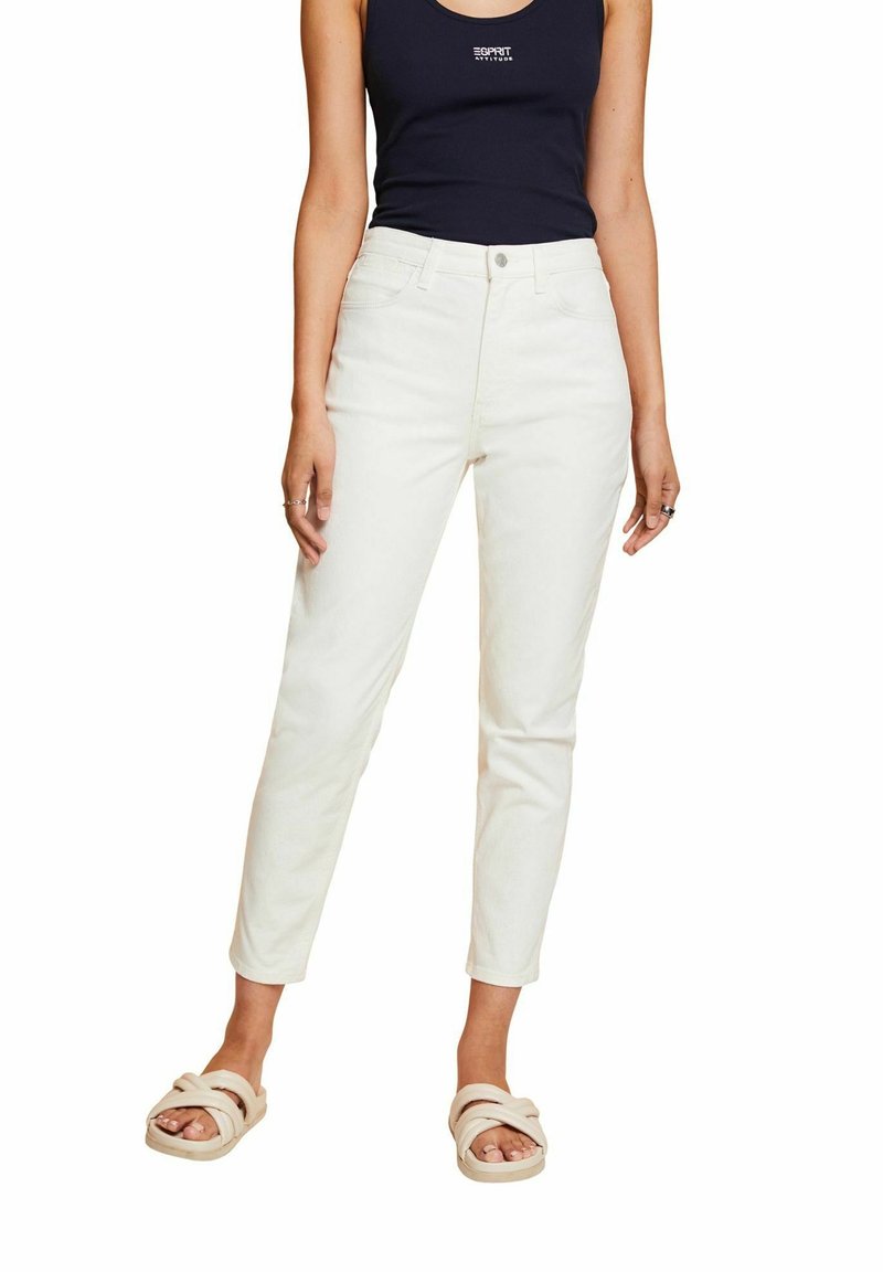 Femme portant un pantalon droit taille haute de couleur blanc cassé, un haut noir sans manches avec texte, et des sandales beiges à enfiler sur fond blanc.