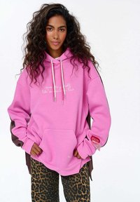 Růžová mikina s kapucí a bílým nápisem, s hnědými akcenty na rukávech. V kombinaci s kalhotami s leopardím vzorem.