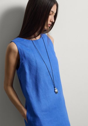 Jonge vrouw met lang donker haar, gekleed in een mouwloze blauwe jurk en een zilveren hanger ketting, staand tegen een grijze muur met handen in de zakken.