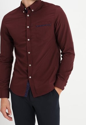 Man wearing a fitted burgundy button-down shirt with a chest pocket and dark navy trousers, one hand in pocket.