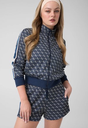 Vrouw draagt een marineblauwe patroon ritsvest en bijpassende korte broek met handen in de zakken, beige hoofdband, lang golvend haar, neutrale gezichtsuitdrukking.