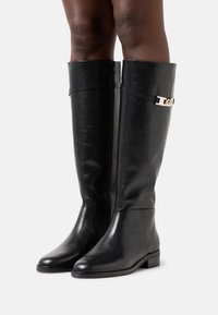 Botas de cuero negras hasta la rodilla con un acabado suave, punta cuadrada, tacón bajo en bloque y un acento metálico en tono dorado cerca de la parte superior.