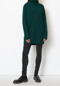 Grüner gerippter Rollkragenpullover mit langen Ärmeln, kombiniert mit schwarzen Skinny-Jeans und klobigen schwarzen Slippern, vor einem weißen Hintergrund.
