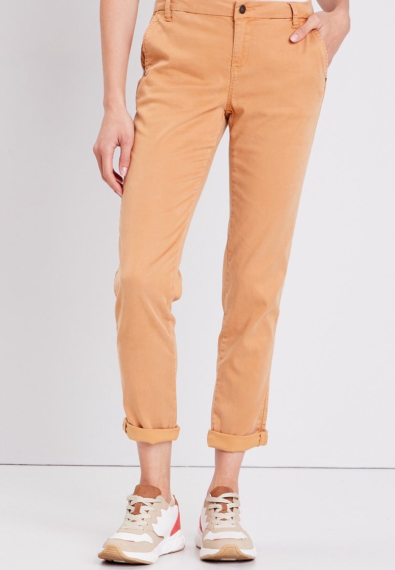 Personne portant un pantalon marron clair retroussé et des baskets beiges avec des touches blanches et corail, debout devant un fond clair uni.