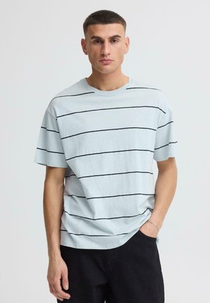 Giovane uomo in piedi con la mano sinistra in tasca, indossa una maglietta azzurro chiaro con sottili strisce orizzontali nere e pantaloni neri, davanti a uno sfondo neutro.