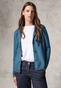 Cardigan teal en tissu tricoté doux, avec une fermeture �à boutons sur le devant, des poignets côtelés et une coupe décontractée. Associé à un t-shirt blanc et à un jean noir.