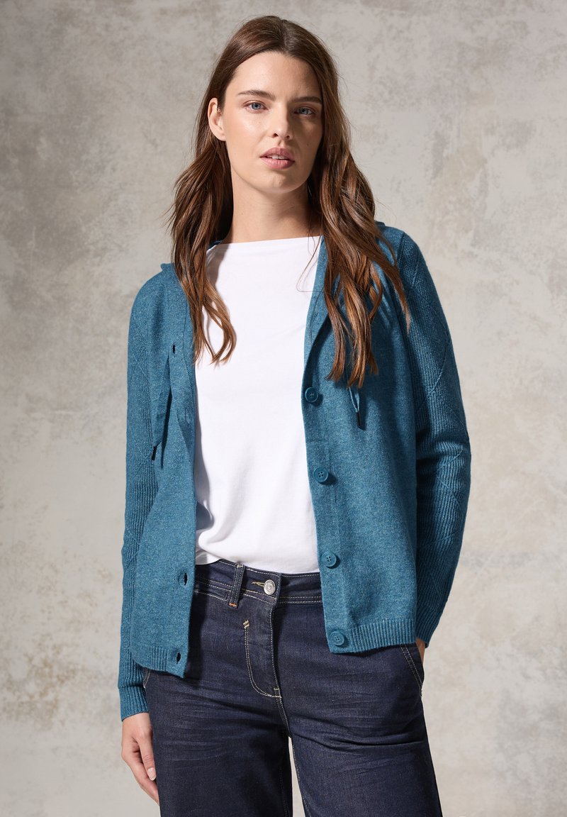 Cardigan teal en tissu tricoté doux, avec une fermeture à boutons sur le devant, des poignets côtelés et une coupe décontractée. Associé à un t-shirt blanc et à un jean noir.