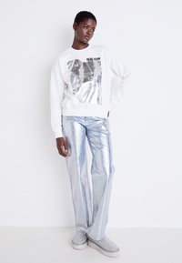 Felpa bianca con stampa grafica, abbinata a pantaloni larghi in metallizzato blu lucido e scarpe slip-on argento scintillanti.