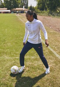 Biela zipsová bunda Puma, námornícké nohavice s ružovými akcentmi a biele športové topánky. Osoba stojí na tráve a ovláda futbalovú loptu.