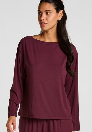 Vrouw met lang donker haar draagt een losse, lange mouwen bordeaux top en bijpassende broek, kijkt iets naar links tegen een effen achtergrond.