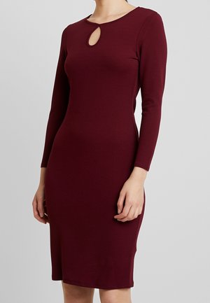 Vrouw draagt een aansluitende, lange mouwen, knielange donkerrode jurk met een kleine druppelvormige uitsparing bij de halslijn tegen een grijze achtergrond.