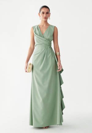 Mujer con vestido cruzado sin mangas color verde salvia, sosteniendo un pequeño bolso beige, accesorios con pendientes y pulseras doradas sobre fondo blanco.