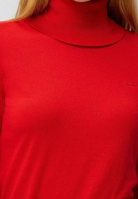 Röd polokragetröja gjord av ett figurnära, slätt material. Har en hög krage och subtil logobroderi på sidan.