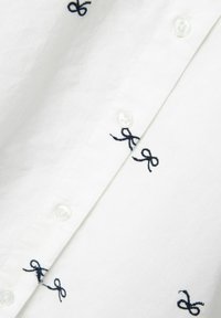 Camisa blanca con patrones de lazo bordados en azul marino, pequeños botones de plástico y una tela de algodón suave con un acabado ligeramente texturizado.