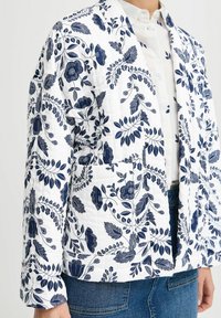 Ciektēta balta jaka ar tumši zilu ziedu rakstu, strukturētu dizainu, garām piedurknēm un priekšējo kabatu. Tekstūrēta auduma ar detalizētu akcentu.
