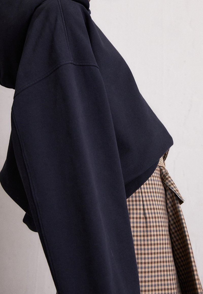 Personne portant un sweat à capuche ample bleu marine foncé assorti à un pantalon à carreaux marron et beige sur un fond clair uni.