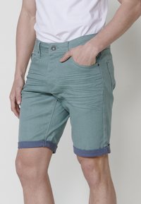 Shorts vert clair avec des revers en bleu foncé, design à cinq poches, fermeture à bouton et tissu texturé. Ajustement slim avec poches latérales.