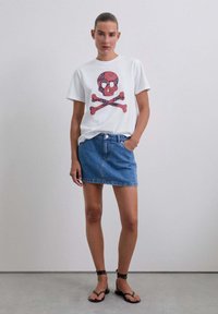 Biała koszulka z czerwono-niebieskim wzorem czaszki i skrzyżowanych kości, zestawiona z dżinsową spódnicą i czarnymi sandałami z ćwiekami.