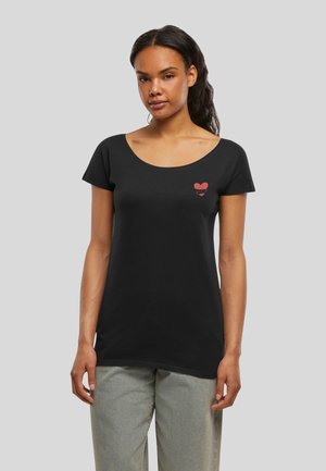 Femme aux cheveux foncés portant un t-shirt noir à manches courtes et encolure arrondie avec un petit motif en forme de cœur rouge sur la poitrine gauche, et un pantalon gris clair.