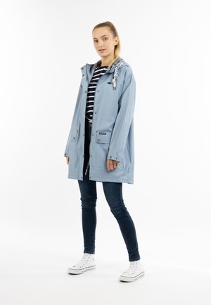 Hellblauer, wasserdichter Mantel mit Reißverschluss, zwei vorderen Taschen und einer gemusterten Kapuze. Getragen über einem schwarz-weißen gestreiften Shirt und blauen Jeans.