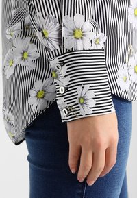 Randig skjorta med blommönster i svart och vitt, med gula accenter, knäppt manschett och mjuk textur. Bärs tillsammans med blå jeans.