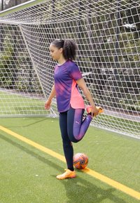 Sportists ar violetā un rozā rakstainu sporta krekls un melnajām legingām izstiepj vienu kāju, uz zaļās zālāja ir oranža futbola bumba.