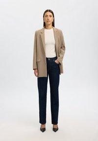 Beige skräddarsydd kavaj, vit T-shirt med rund halsringning, mörkblå raka jeans och svarta spetsiga klackar. Modellen står mot en ljus bakgrund.