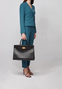 Svart läderhandväska med strukturerad design, guldiga detaljer och överhandtag, hålls bredvid en person i en teal kostym och högklackade sandaler.