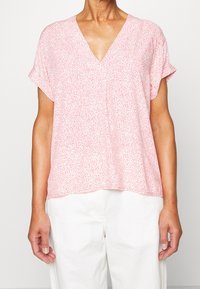 Rosa kurzärmlige Bluse mit V-Ausschnitt und einem strukturierten Muster, das kleinen Punkten ähnelt. Kombiniert mit hellen Hosen.