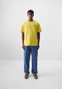 Camiseta de algodón amarilla con cuello redondo, mangas cortas y gráficos verdes. Combinada con unos vaqueros azules y zapatos marrones.