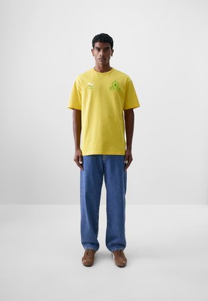 Camiseta de algodón amarilla con cuello redondo, mangas cortas y gráficos verdes. Combinada con unos vaqueros azules y zapatos marrones.