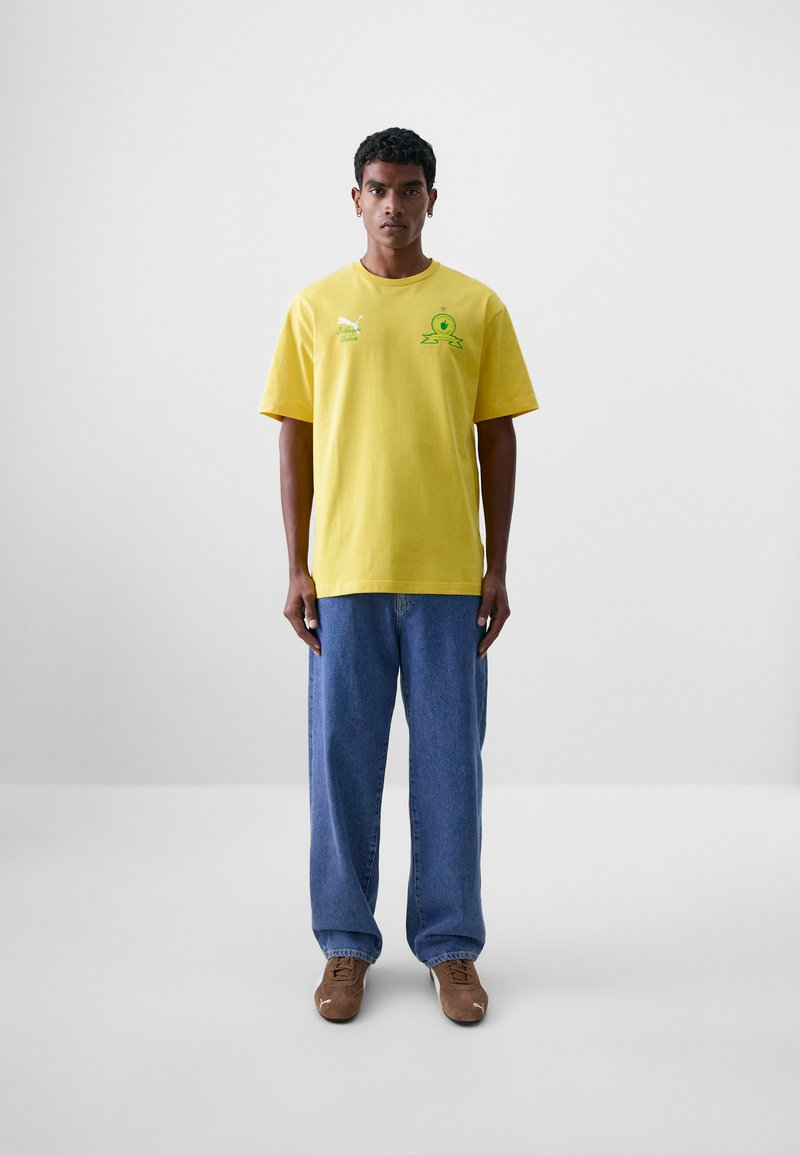 Camiseta de algodón amarilla con cuello redondo, mangas cortas y gráficos verdes. Combinada con unos vaqueros azules y zapatos marrones.