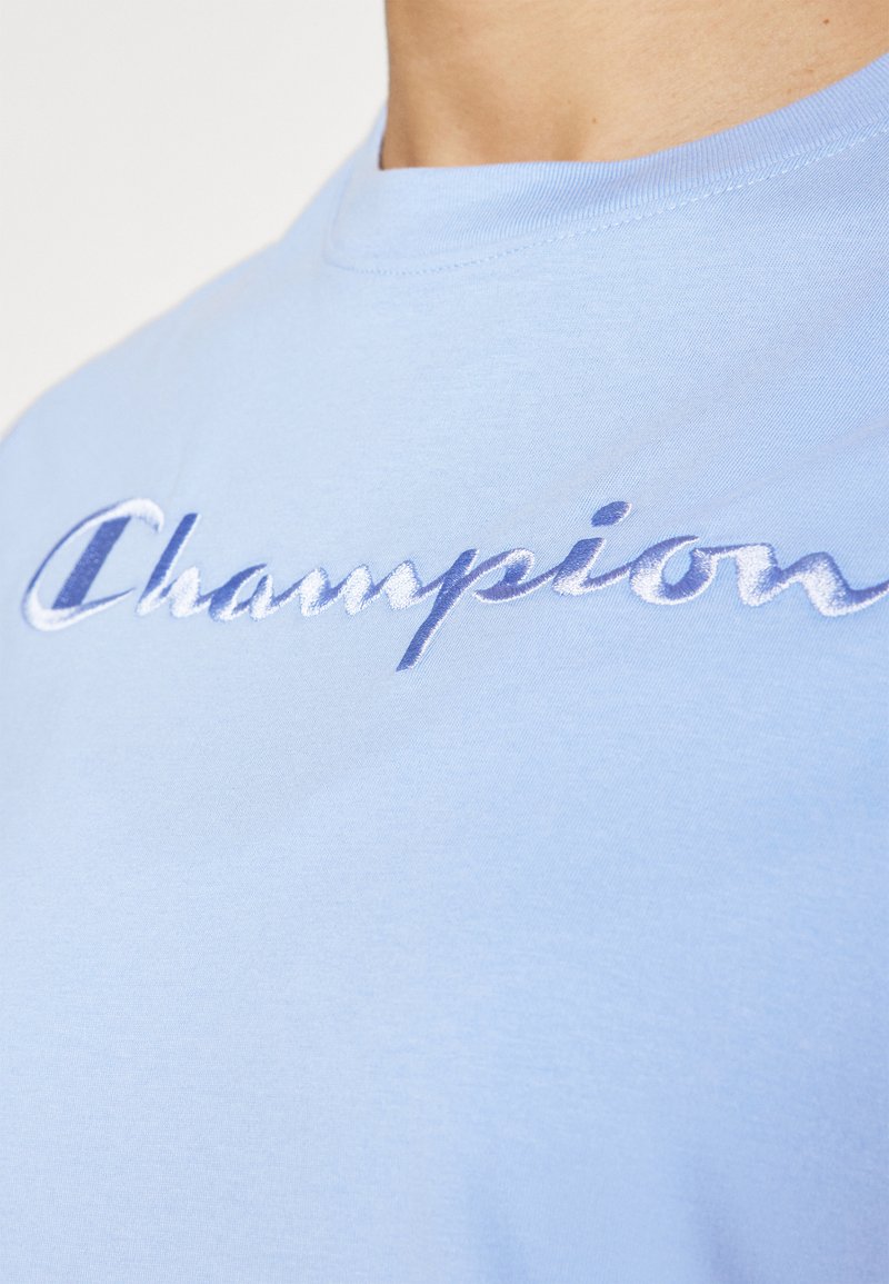 T-shirt a girocollo in cotone azzurro chiaro con logo "Champion" ricamato in blu scuro, texture liscia e maniche corte.