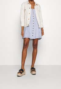 Veste en denim blanc portée sur une robe d'été rayée bleu et blanc avec une patte de boutonnage à l'avant, associée à des sandales à plateforme noires avec une semelle texturée.