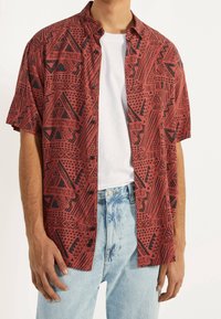 Camisa roja de manga corta con botones y patrones geométricos negros, llevada sobre una camiseta blanca, combinada con jeans de mezclilla azul claro.