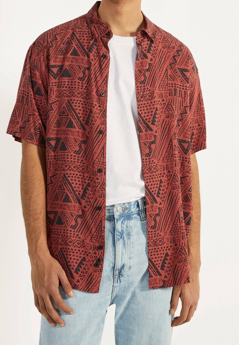 Camisa roja de manga corta con botones y patrones geométricos negros, llevada sobre una camiseta blanca, combinada con jeans de mezclilla azul claro.