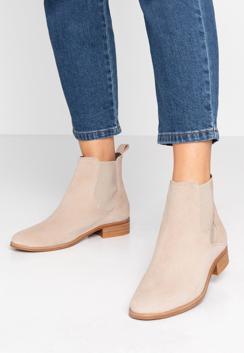 Bottes cheville en daim beige avec talons bas en bois portées avec un jean en denim bleu coupé au-dessus de la cheville sur un sol blanc.