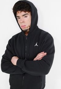 Musthave musta kapuutsiga sviiter, valmistatud pehmest kangast, millel on ees tasku. Vasakul rinnal on valge Jumpmani logo ja reguleeritav kapuuts.