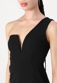 Svart jumpsuit med dyp V-hals, asymmetrisk design med én skulder, tettsittende bodice og strukturert midje. Glatt, teksturert stoff.
