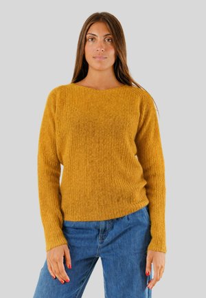 Donna con capelli castani lunghi che indossa un maglione lavorato a maglia di colore giallo senape e jeans blu, in piedi davanti a uno sfondo grigio chiaro uniforme.
