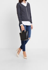 Grå tröja med V-ringning och nitsmyckningar, lager-på-lager över en vit skjorta. Mörka jeans med trasiga knän och beiga högklackade skor. Liten svart handväska.